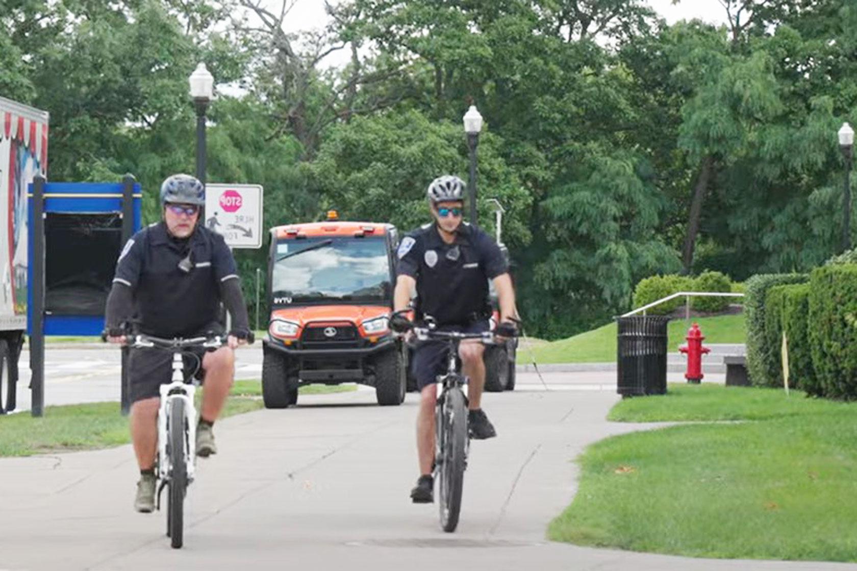 wo uniformed officers on bicycles ride towards the camera on a paved path, with an orange utility vehicle behind them and green trees in the background.